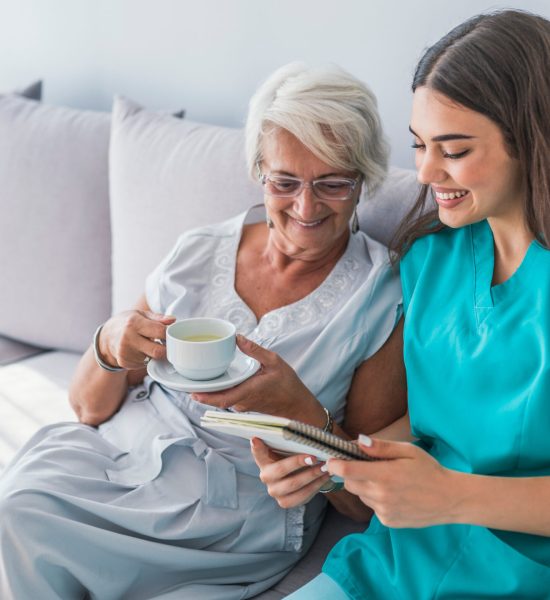 Woman reading a book while sitting with happy grandmother drinking tea. Happy elder woman sitting on white sofa and listening to nurse reading a book out loud Woman reading a book while sitting with happy grandmother drinking tea. Happy elder woman sitting on white sofa and listening to nurse reading a book out loud